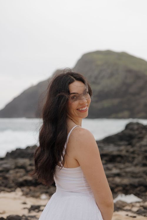 Brunette woman in white dress smiling over shoulder on rocky beach with green headland behind her.