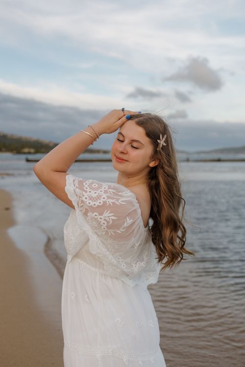 A young woman in a white lace dress stands on a beach at sunset with a starfish hair accessory.