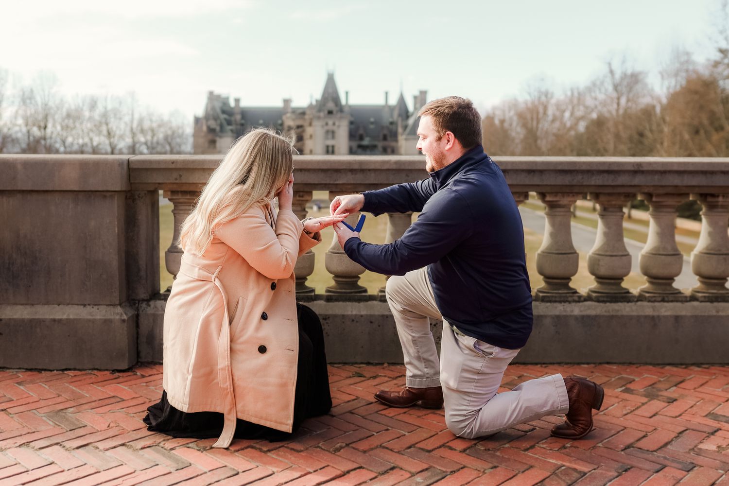 Man proposes to woman on one knee on brick terrace with historic Biltmore Estate in background.