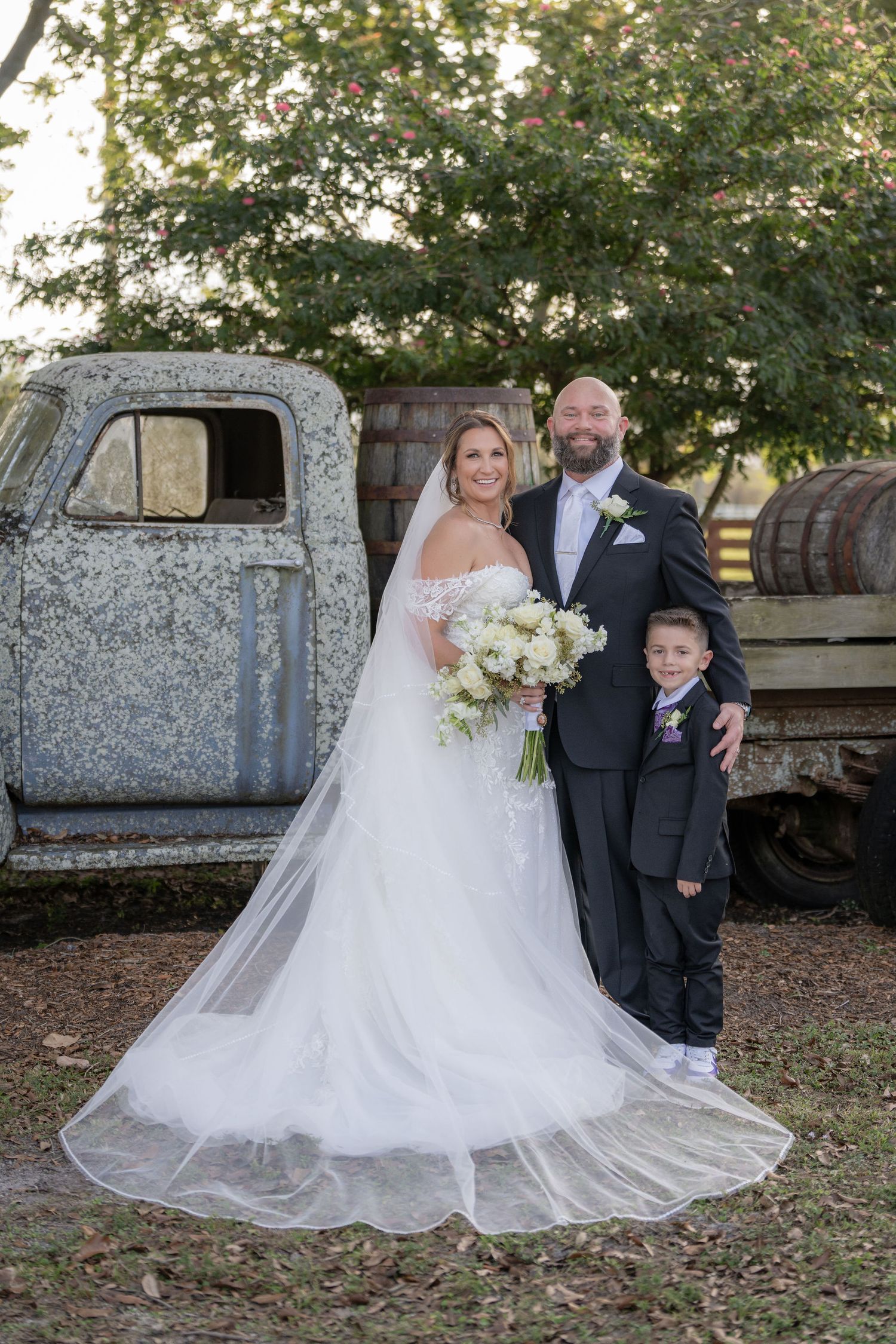 Bride, groom, and son posing by the vintage farm truck and barrels at a Rockin' H Ranch wedding in Palm City, FL