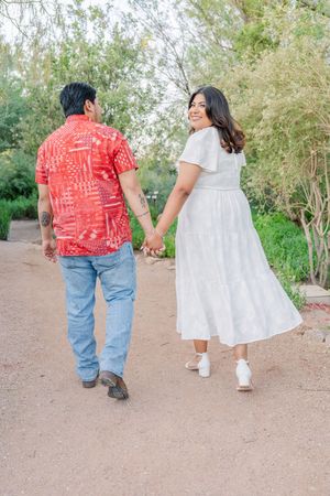 Couple holding hands while walking in a garden, woman in white dress looking back smiling.