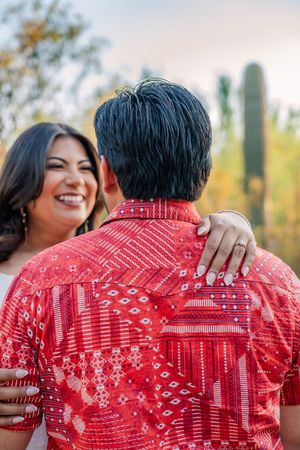 Smiling woman embraces man in red patterned shirt outdoors near desert cacti.
