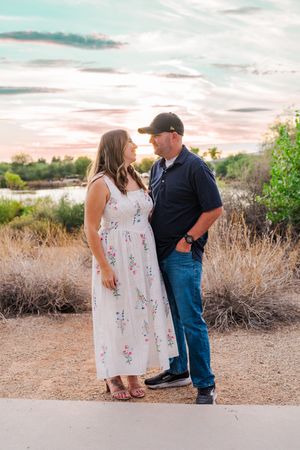 Couple standing face to face on a path at sunset, woman in floral maxi dress and man in navy polo and cap.