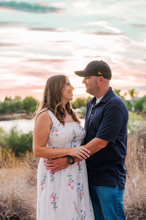 Couple holding each other in a field at sunset, woman in floral dress and man in navy polo and black cap.