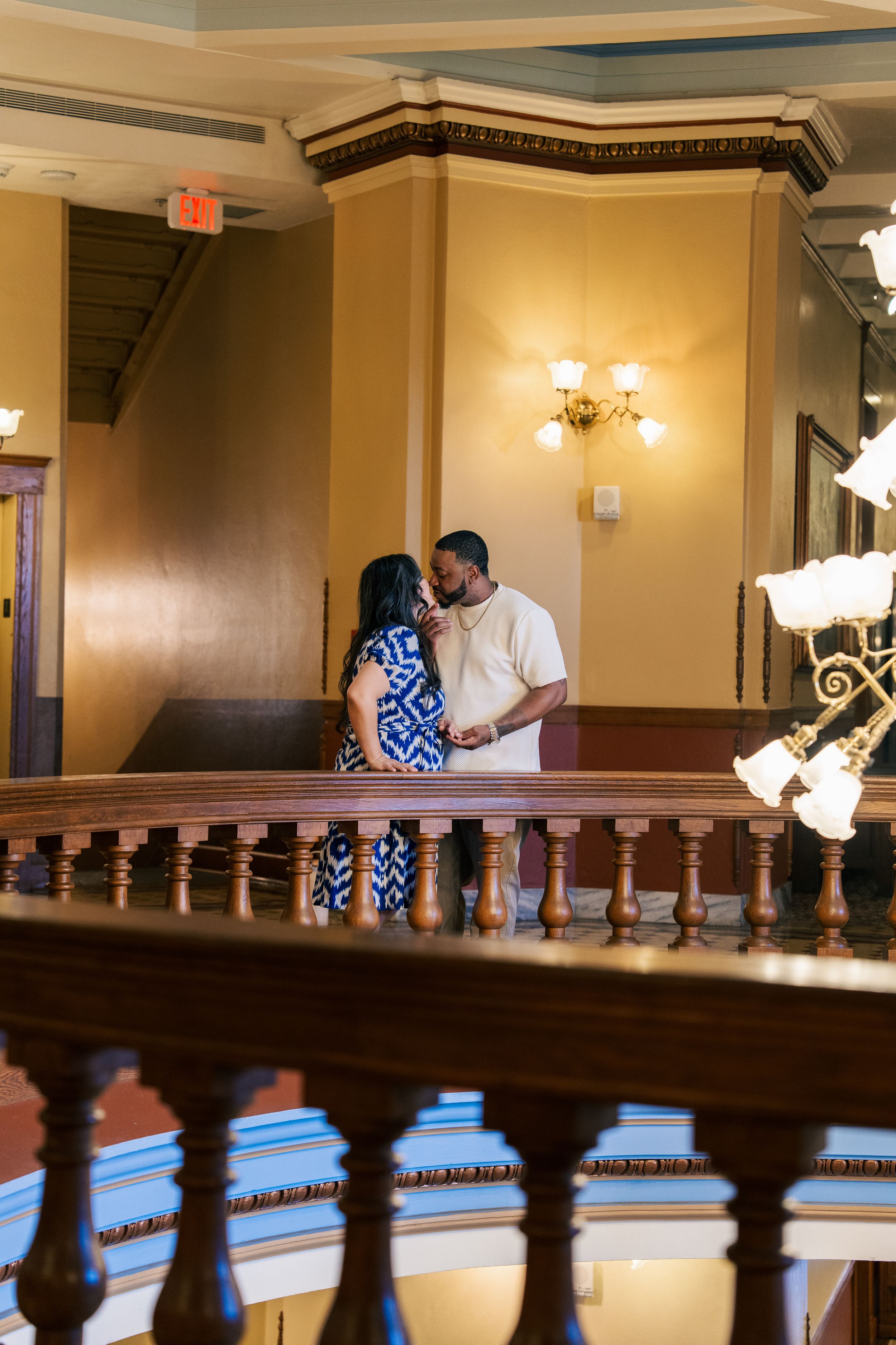 Couple sharing an intimate moment on a balcony inside an elegant historic building with chandeliers.
