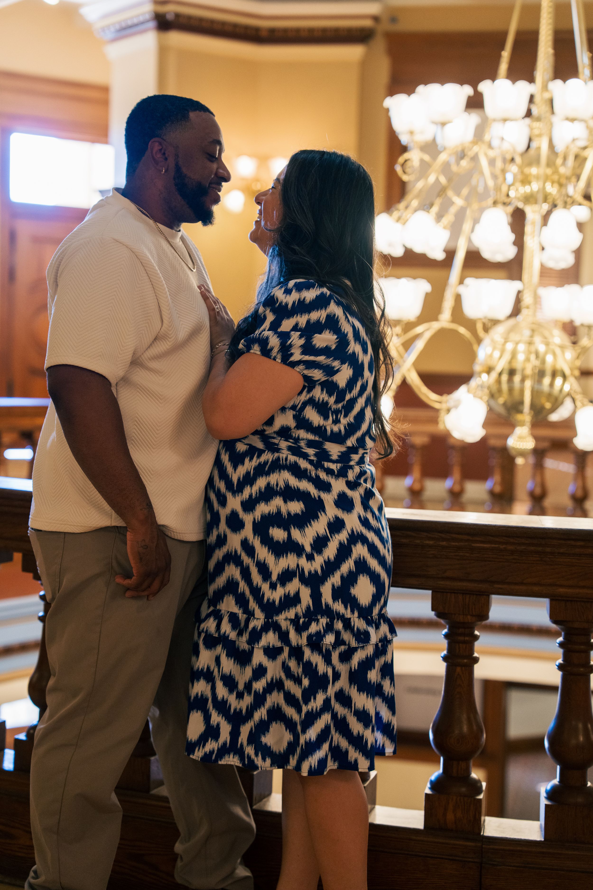 Couple gazing at each other lovingly near wooden railing inside elegant historic building with chandelier.