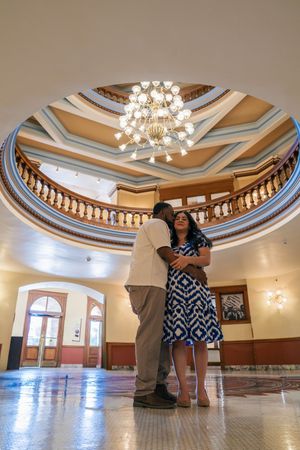 Couple embracing in grand rotunda of historic building with ornate chandelier and circular balcony above.