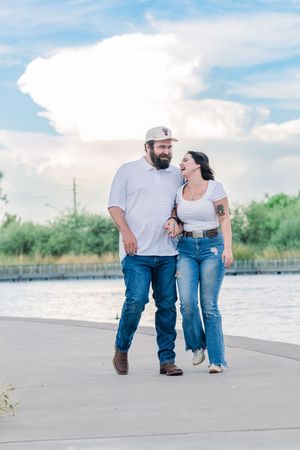 Happy couple walking hand-in-hand along a waterfront path under a dramatic cloudy sky.