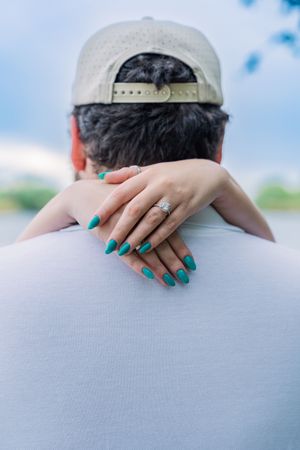 Close-up of womans hand with engagement ring and teal nails resting on mans shoulder from behind.