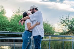 Couple sharing a kiss while embracing near a fence with lush green trees in the background.