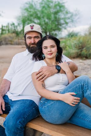 Couple sitting together on a bench outdoors, man with beard in white shirt and cap embracing dark-haired woman.