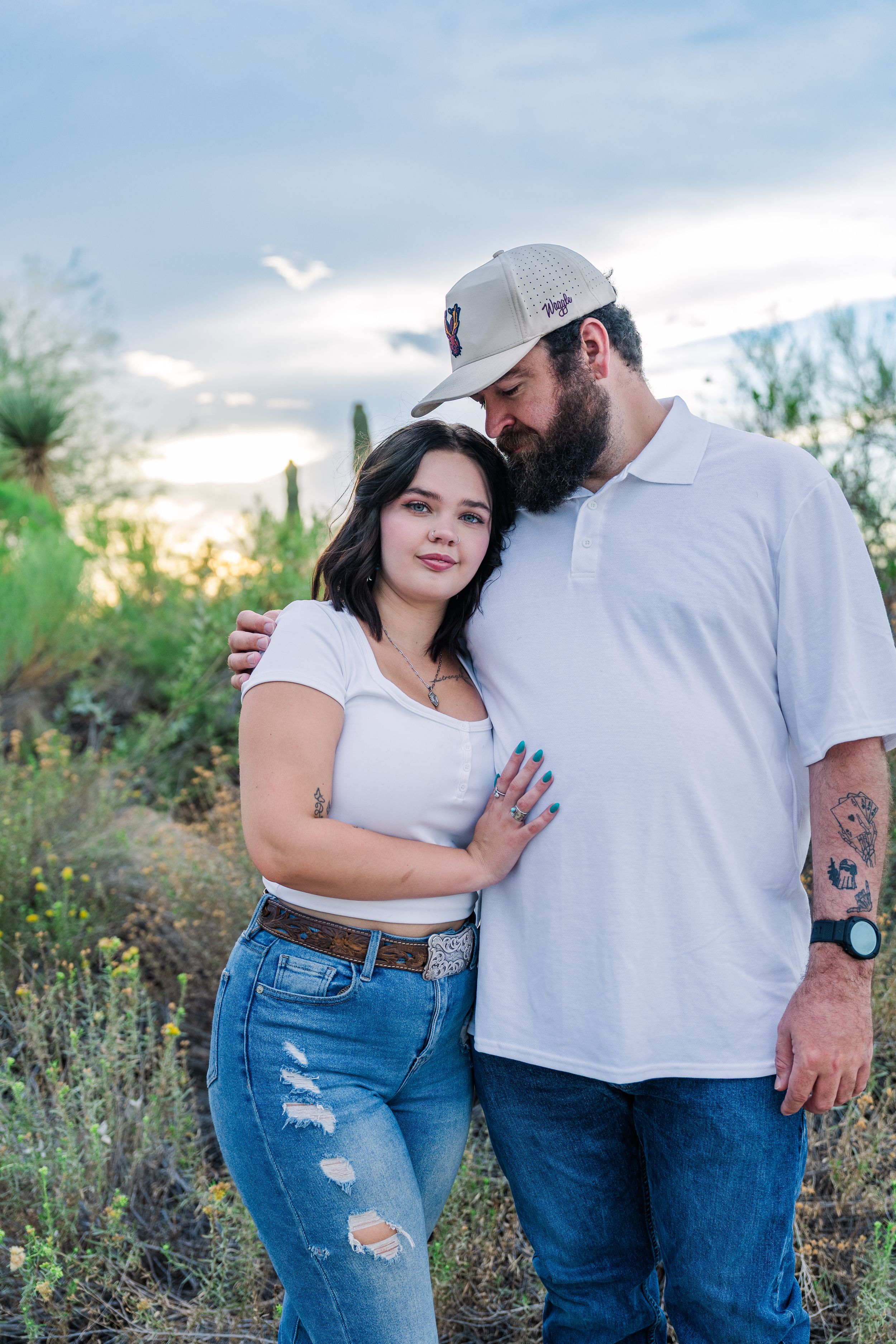 Couple embracing in desert landscape with saguaro cacti, woman in white crop top and jeans, man in white polo and cap.