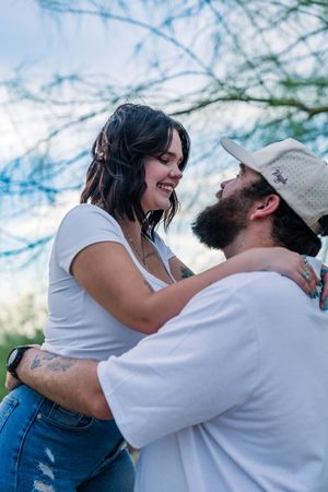 A woman smiles up at a bearded man in a cap as they embrace closely under a blue cloudy sky.