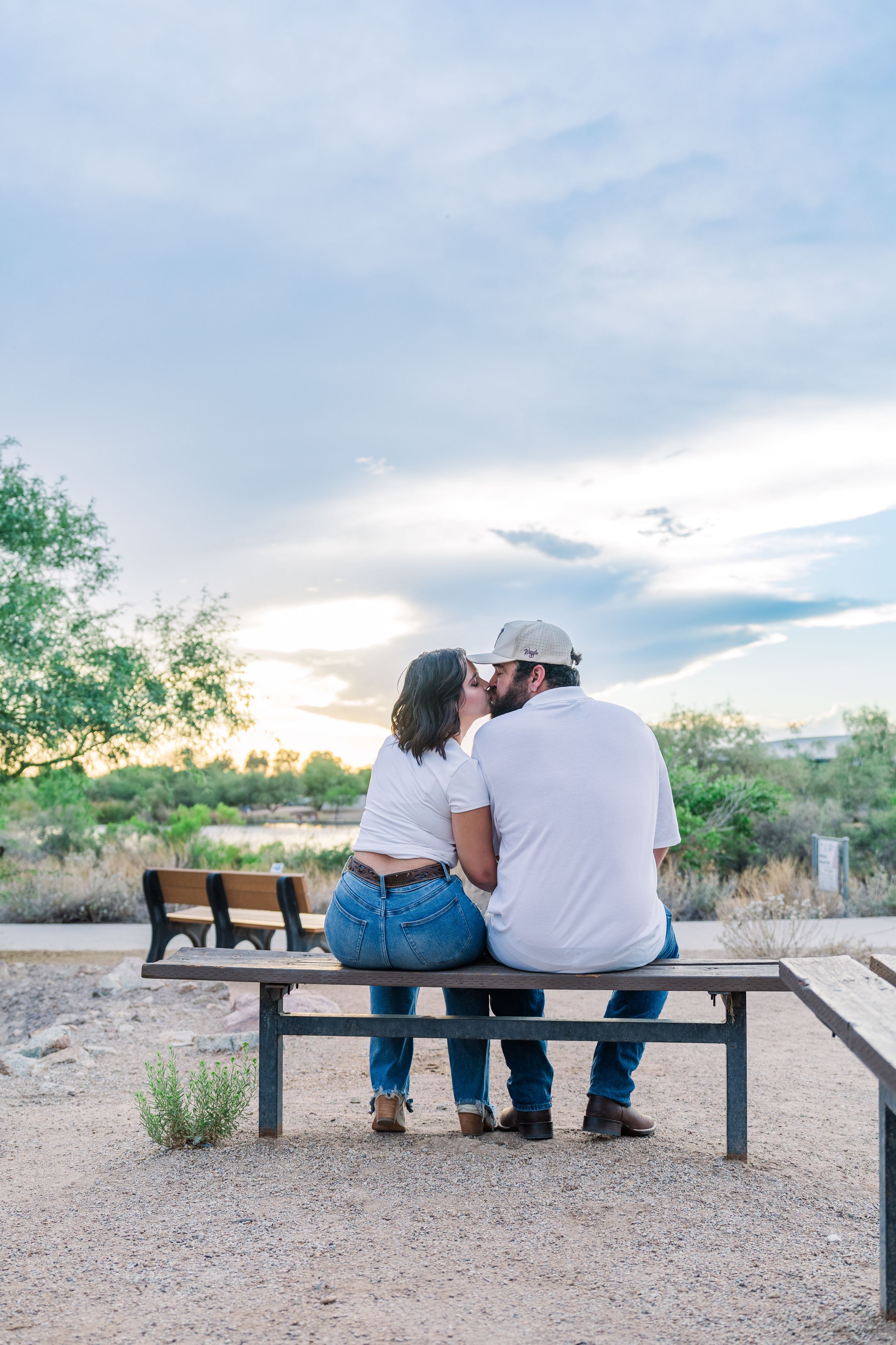A couple shares a kiss while sitting together on a bench at sunset in a desert park setting.