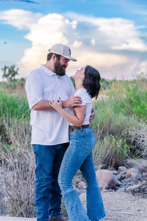 A bearded man in a cap and white shirt embraces a woman in jeans against a scenic desert landscape.