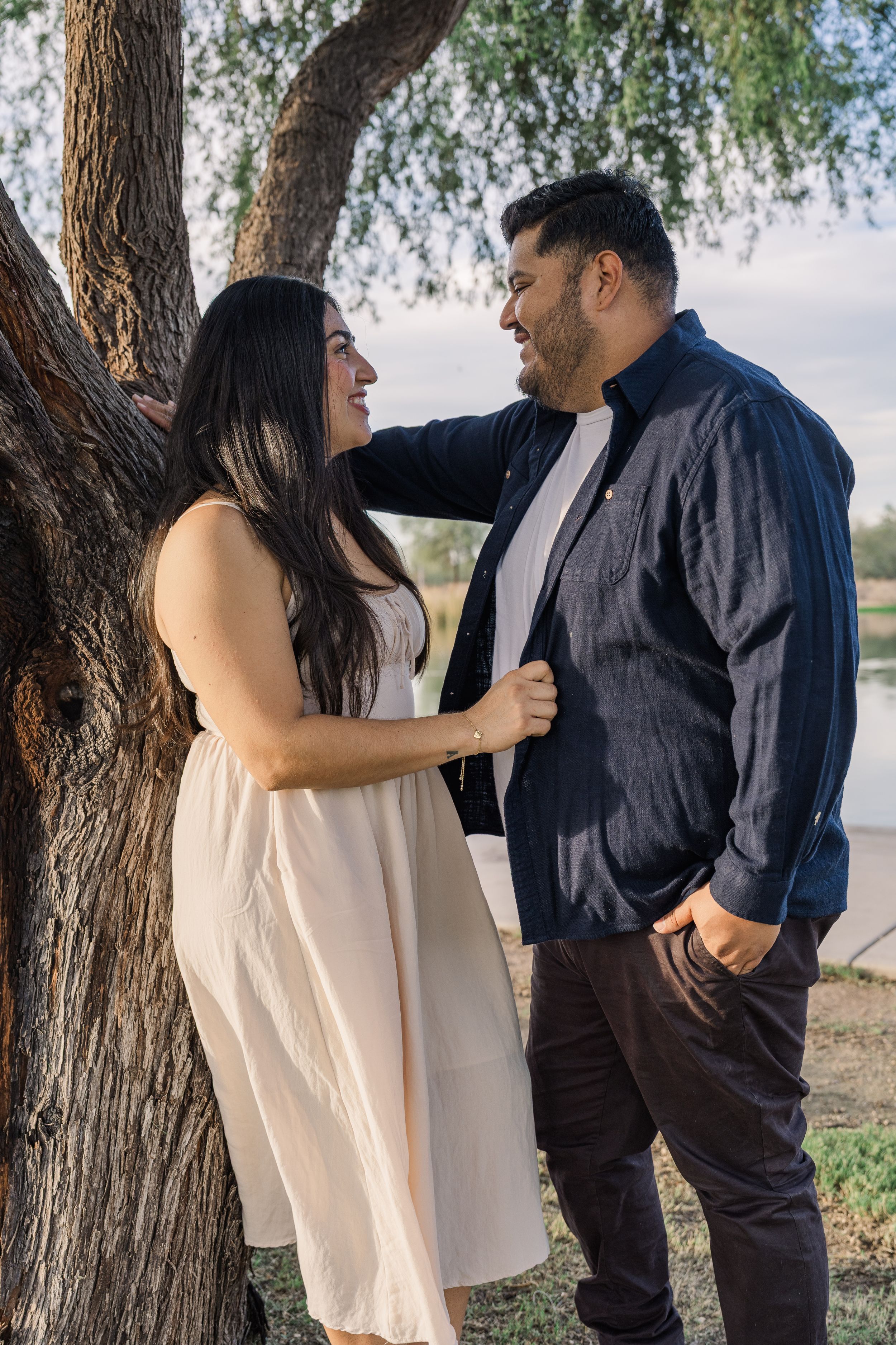 A smiling couple leans against a large tree trunk near a lake, gazing at each other affectionately.