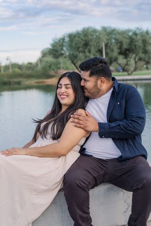 A couple sits together on a ledge by a lake, the man embracing the laughing woman from behind.