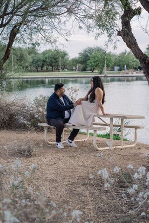 A couple holds hands while sitting at a picnic table near a tranquil lake surrounded by bare trees.