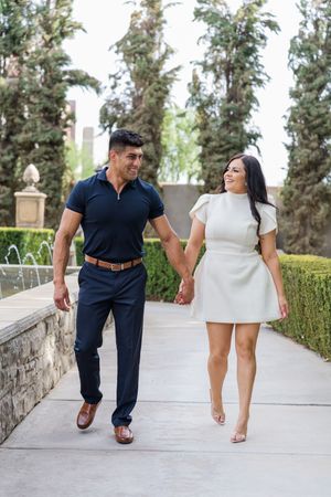 A happy couple holds hands and smiles at each other while walking along a garden pathway with fountains.