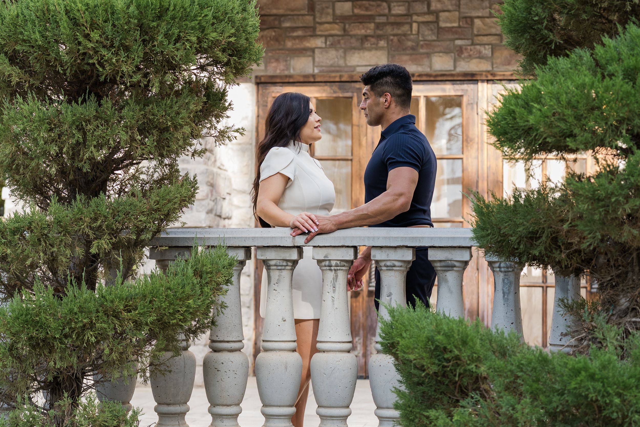 A couple gazes into each others eyes on a balcony surrounded by manicured pine trees and a stone building.