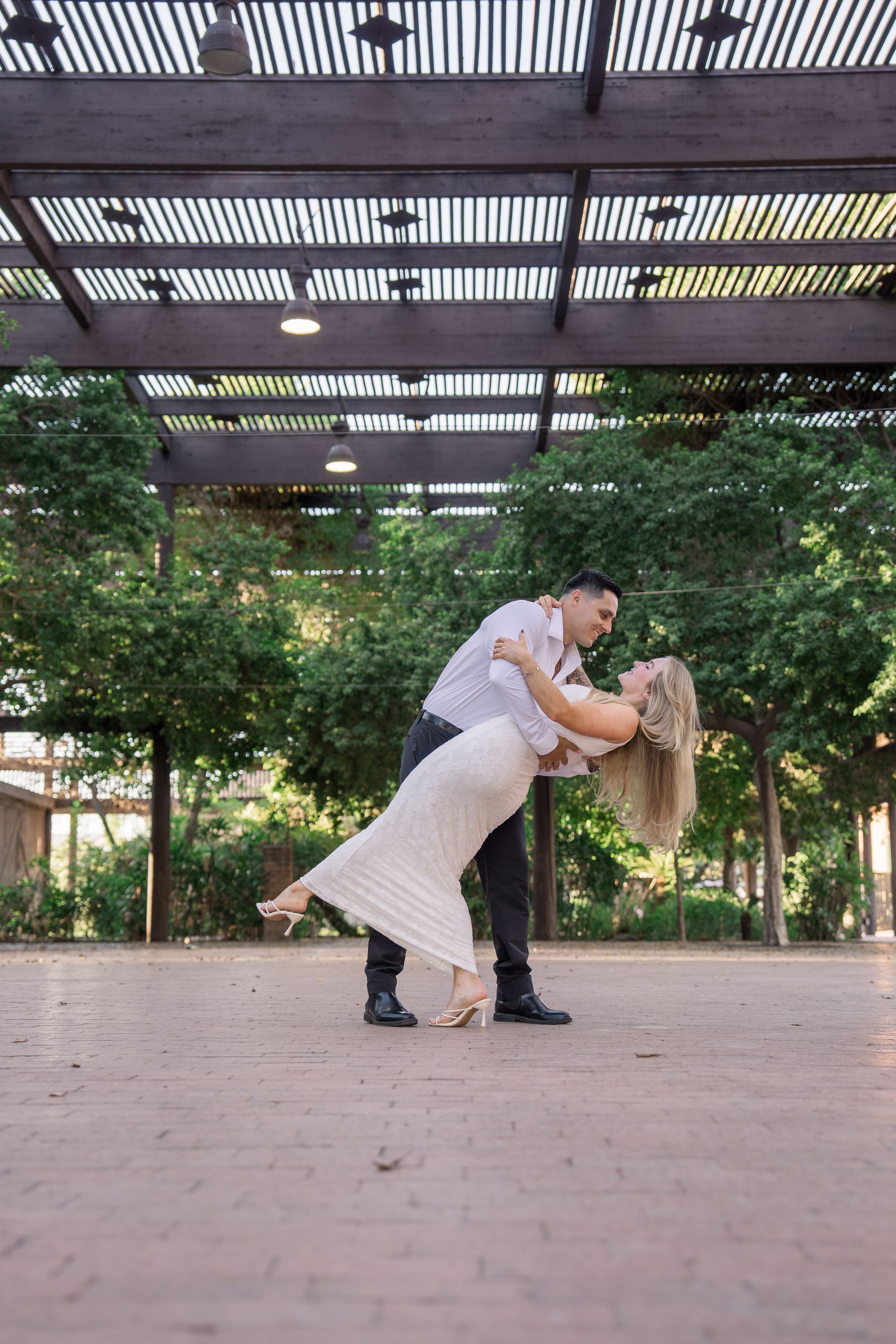 Groom dips blonde bride in romantic dance pose under wooden pergola in garden setting.