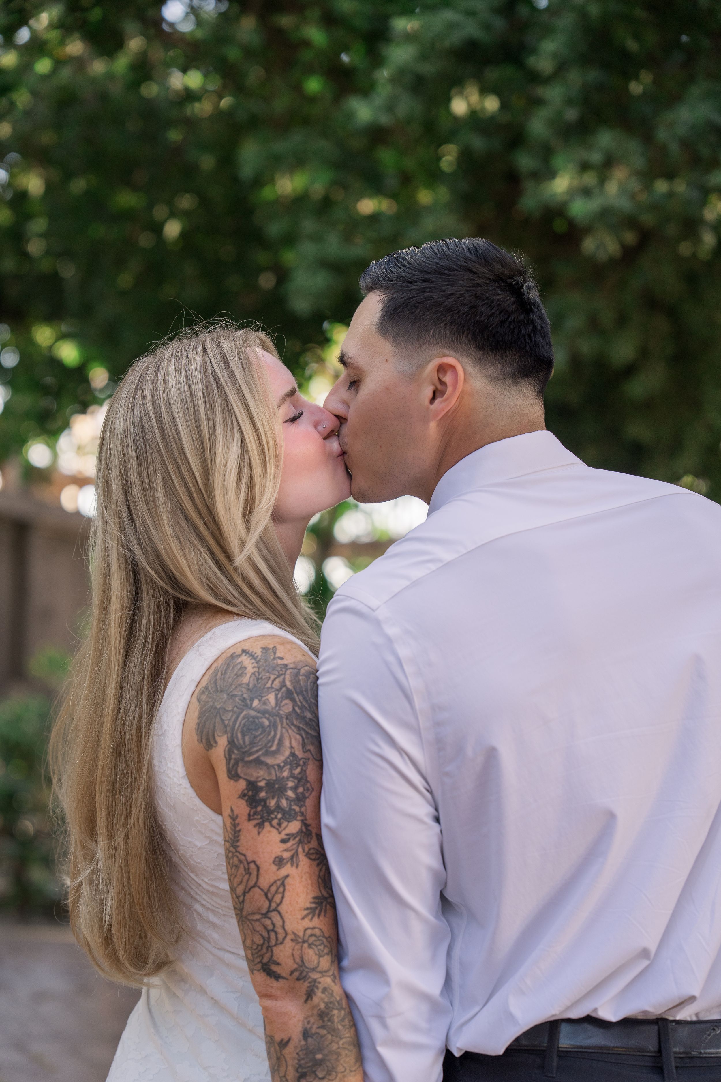 Tattooed blonde bride and groom share passionate kiss in outdoor garden setting.