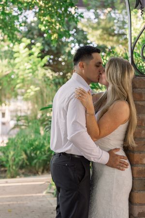 Couple shares romantic kiss against brick wall surrounded by lush green garden foliage.