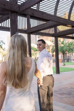 Smiling groom in white shirt turns to hold hands with blonde bride under wooden pergola.