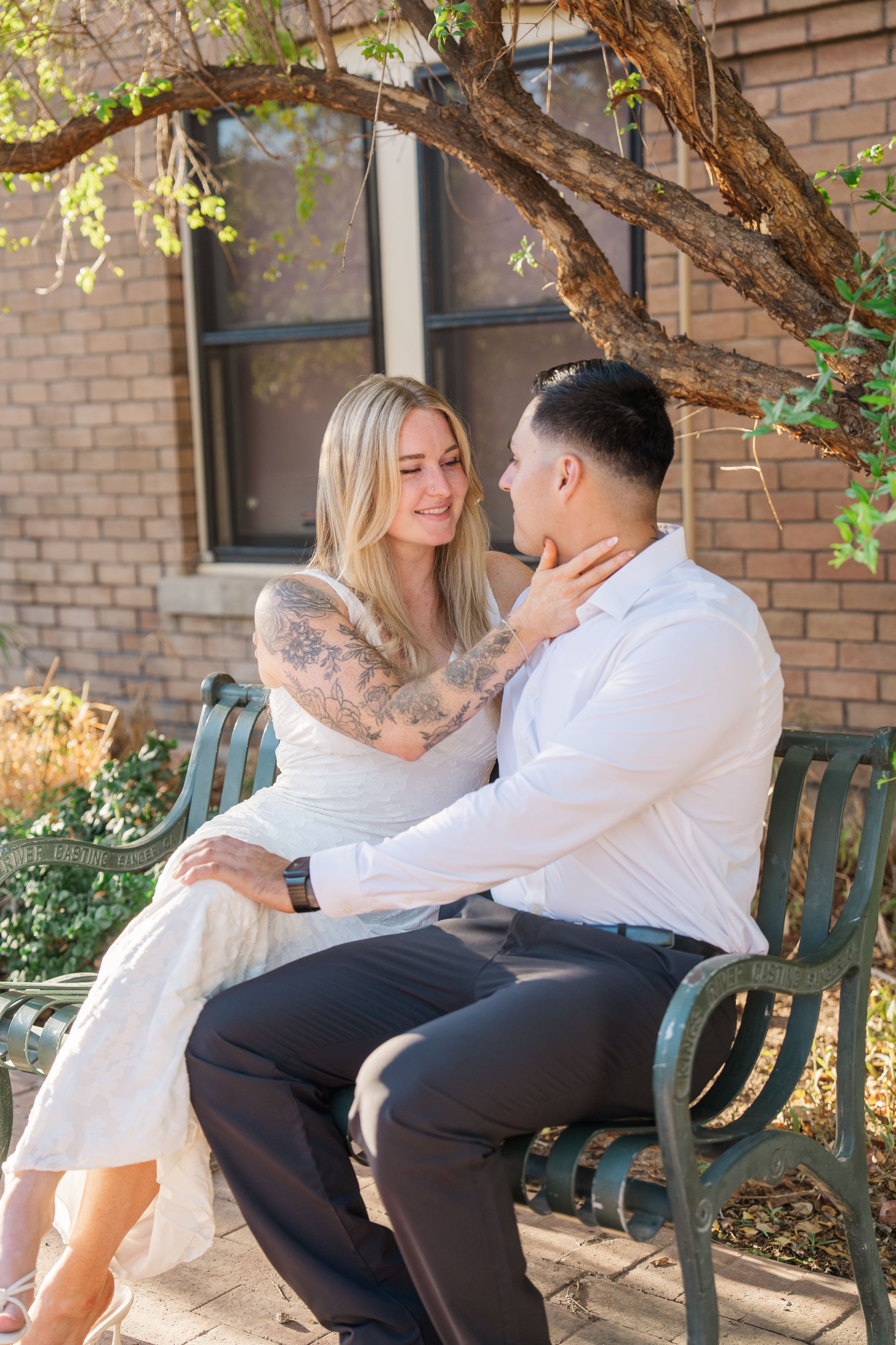 Couple sits on garden bench as tattooed blonde bride tenderly touches grooms face outdoors.