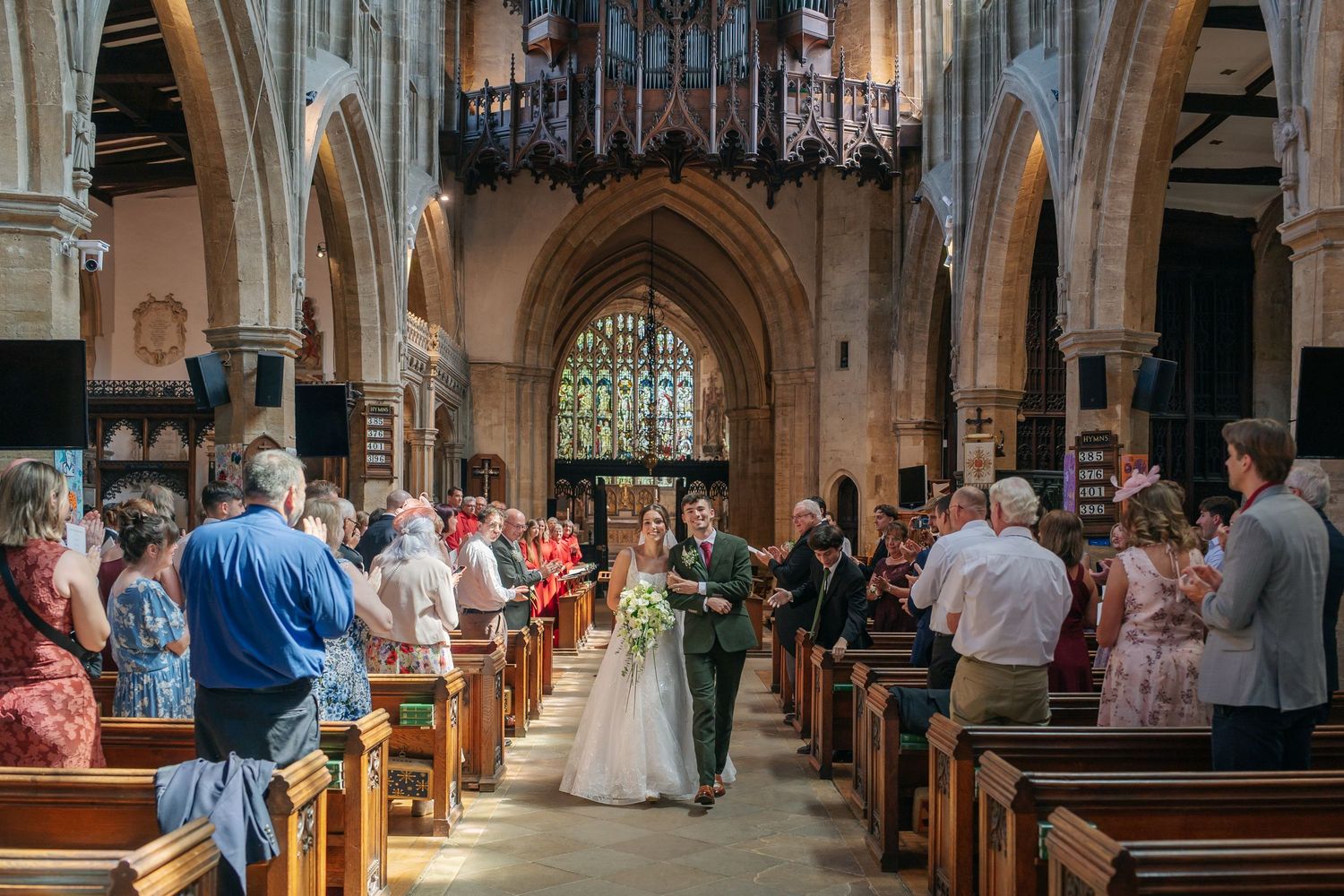 Bride and groom walk down the aisle of a stunning Gothic church, surrounded by standing wedding guests.