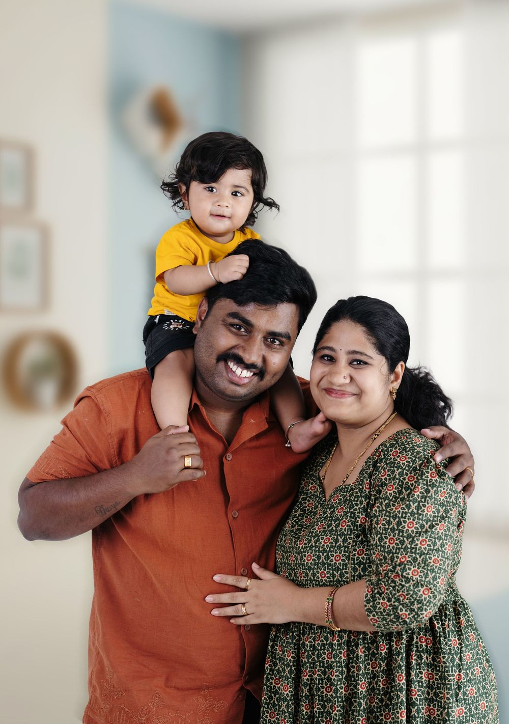 Happy Indian family portrait with father, mother, and toddler child sitting on dad's shoulders indoors.