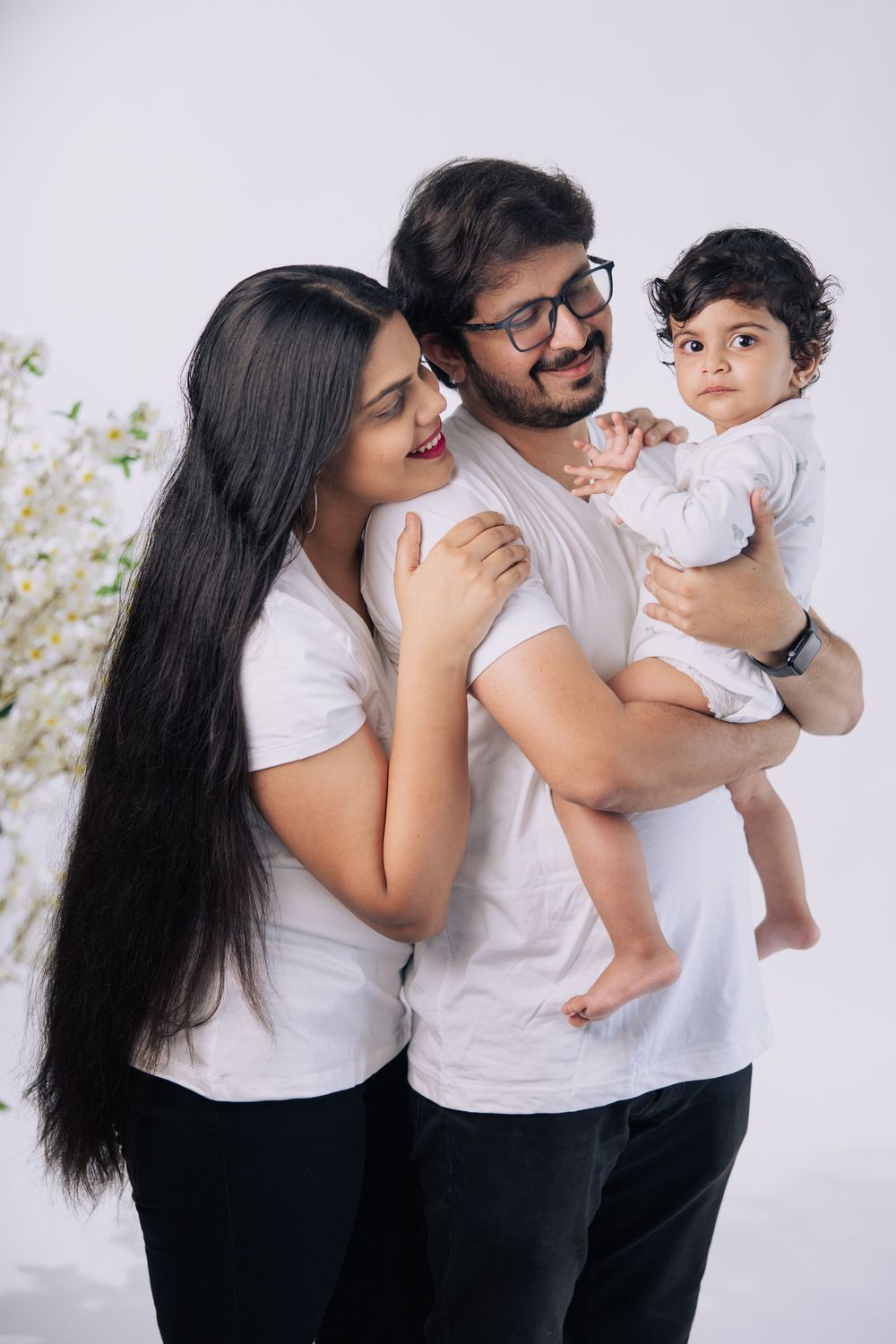 Happy Indian family portrait with mother, father in glasses, and baby in white outfits on light background.
