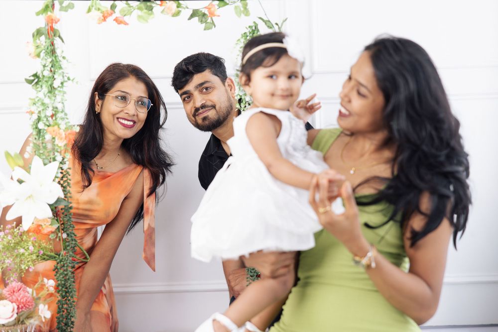Family celebration with adults and baby girl in white dress, posing before floral backdrop at festive event.