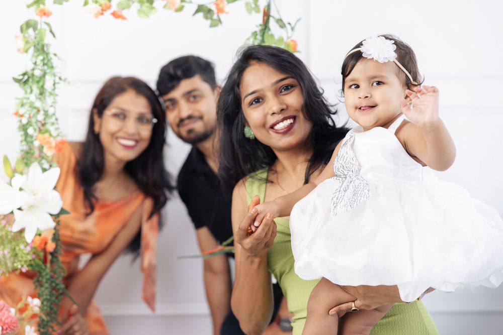 Happy Indian family posing for a portrait with a baby girl in a white dress, smiling in a decorated studio setting.