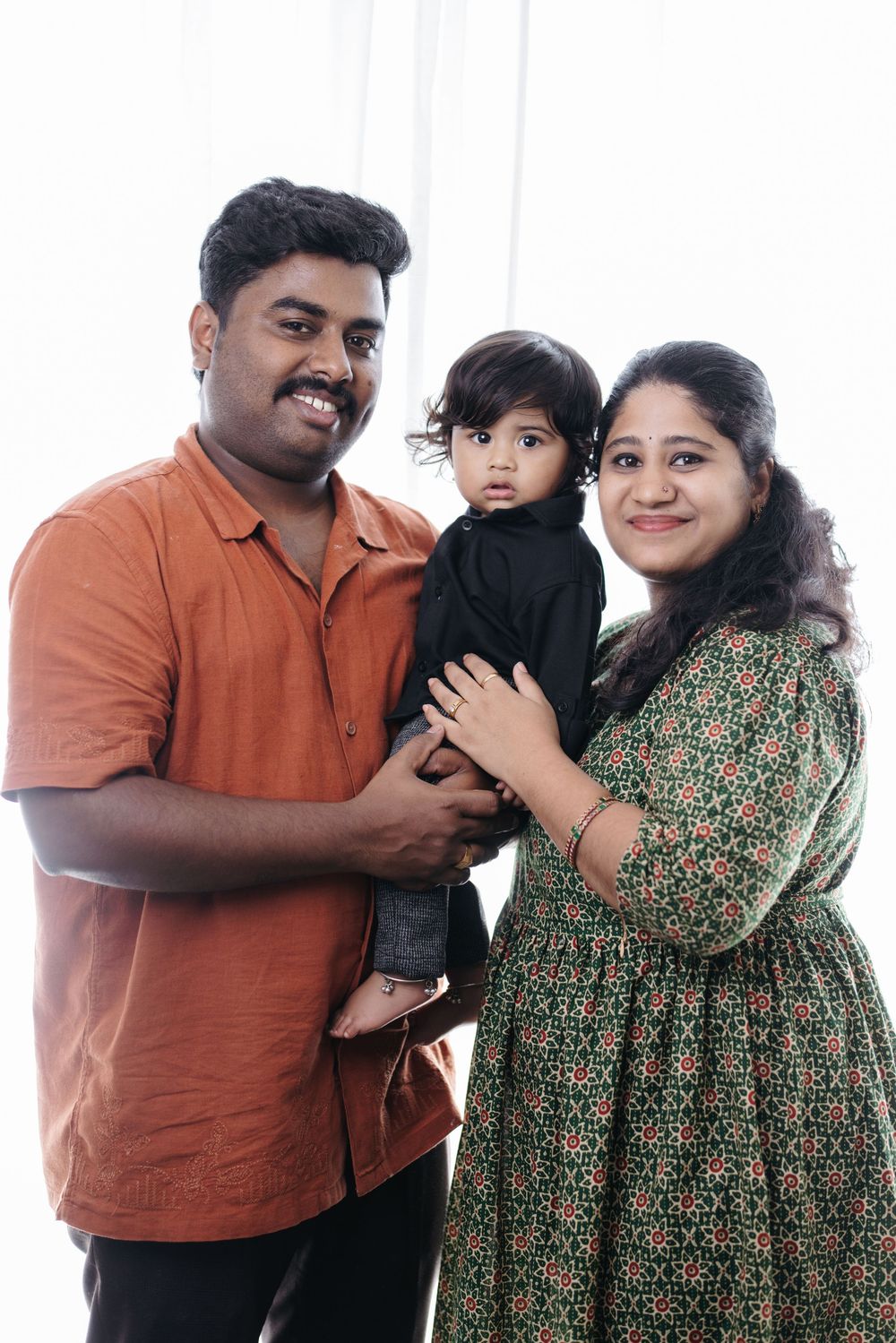 Happy Indian family portrait with father in orange shirt, mother in green dress, holding toddler in black outfit.