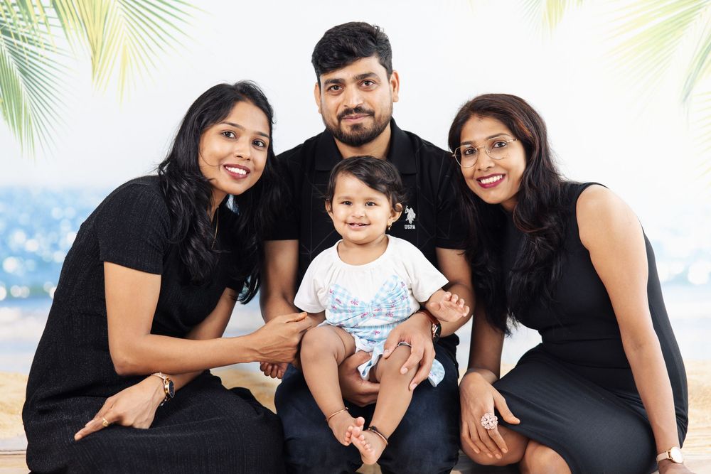 A joyful South Asian family of three adults in black outfits posing with a smiling baby in a bright studio setting.