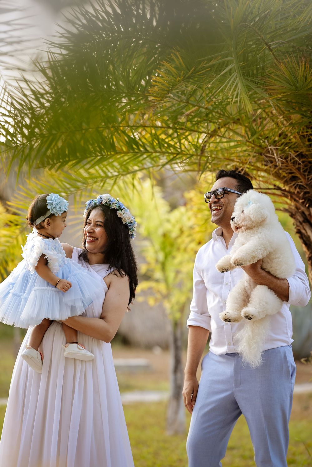 Happy family with toddler girl in blue dress and white fluffy dog posing outdoors near tropical palm trees.