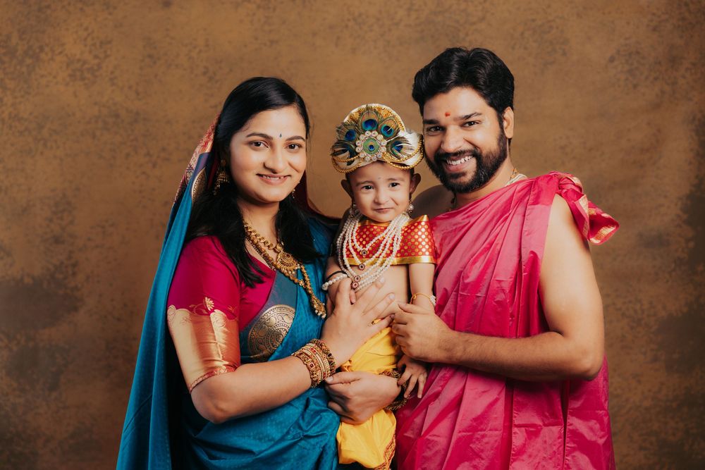 Indian family in traditional attire, parents in saree and dhoti holding child dressed as Lord Krishna.