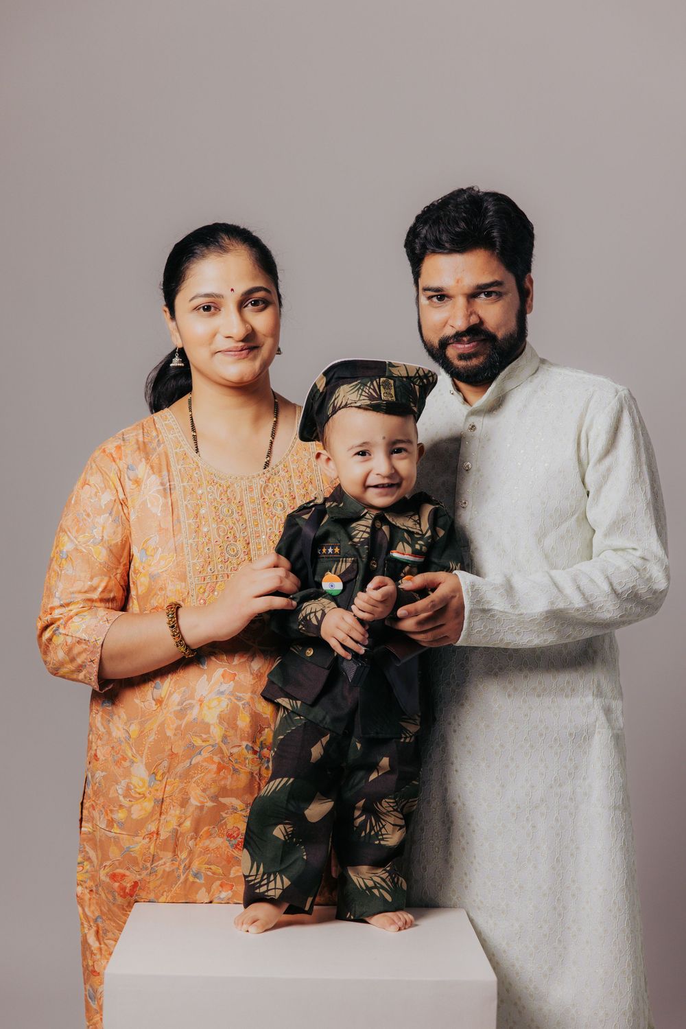 Indian family portrait with mother in orange salwar, father in white kurta, and baby in traditional outfit.