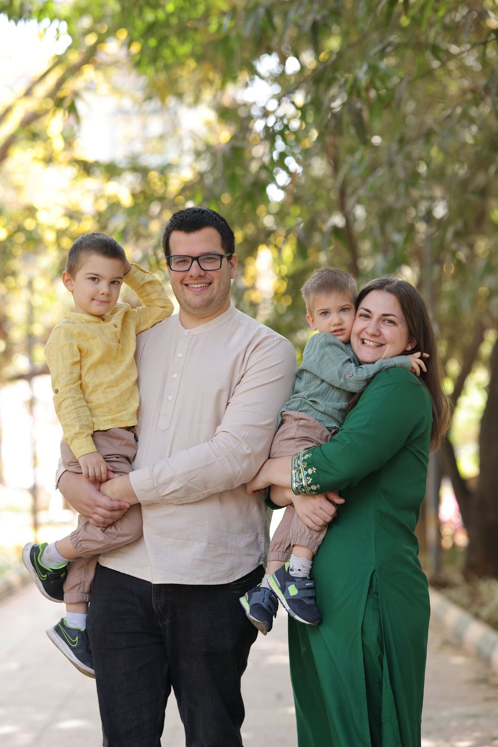 A smiling family of four poses outdoors, with parents holding two young boys, surrounded by lush green trees.