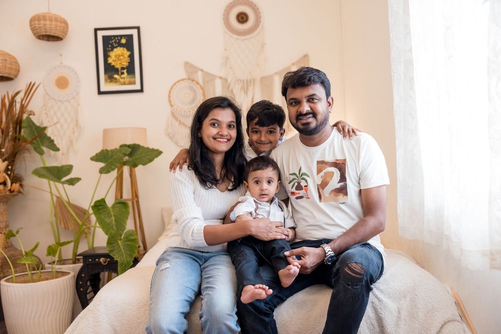 A happy South Asian family of four poses together on a couch in a cozy boho-styled living room.