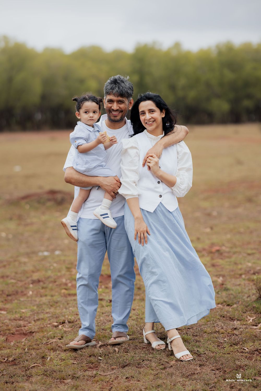 A happy family of three poses outdoors, parents in white and blue outfits holding their toddler in a scenic field.