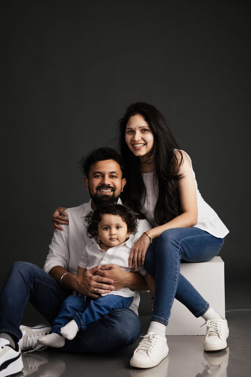 A happy South Asian family of three poses for a studio portrait, parents smiling with their curly-haired toddler.