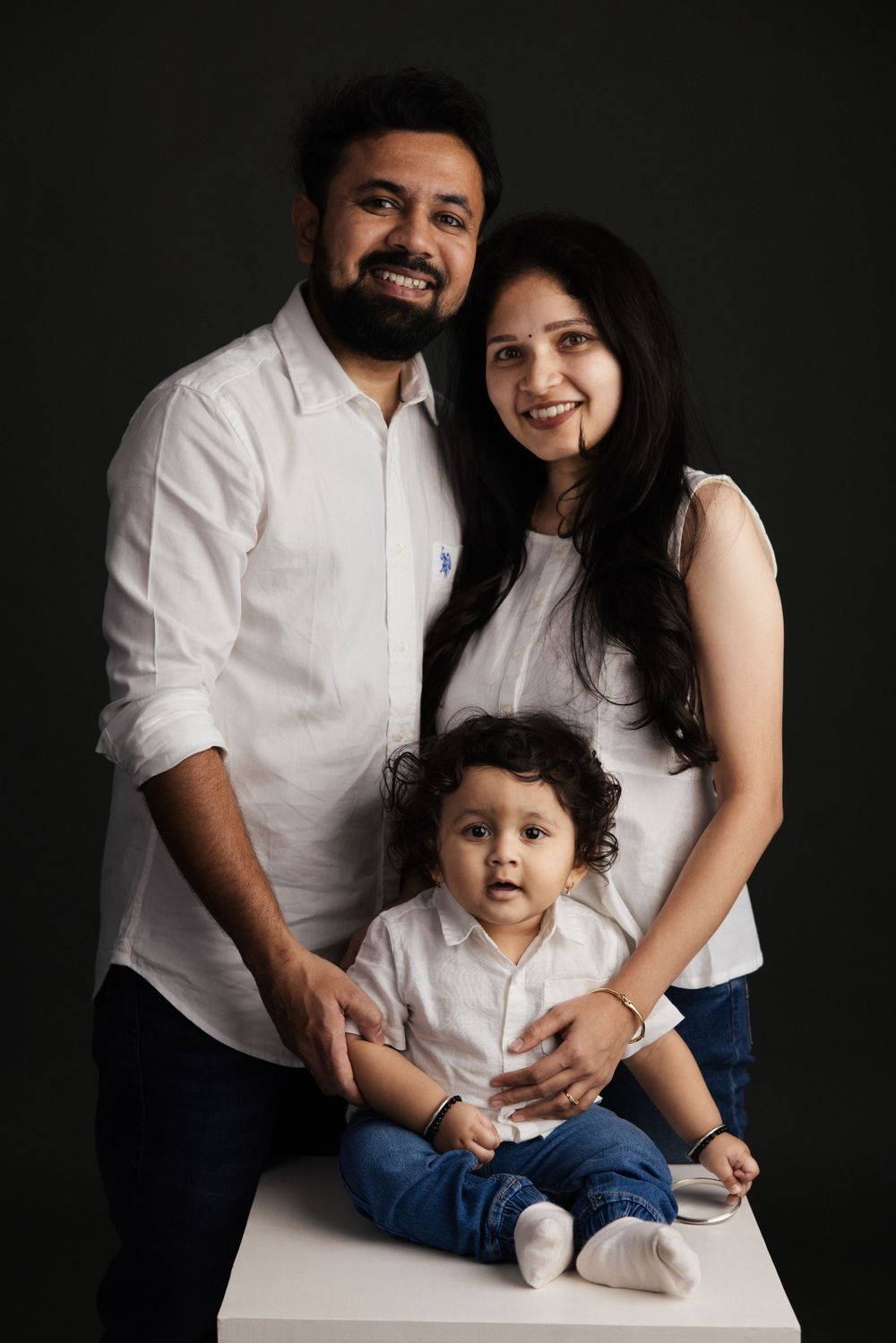 A happy Indian family of three in white shirts and jeans poses for a studio portrait against a dark background.