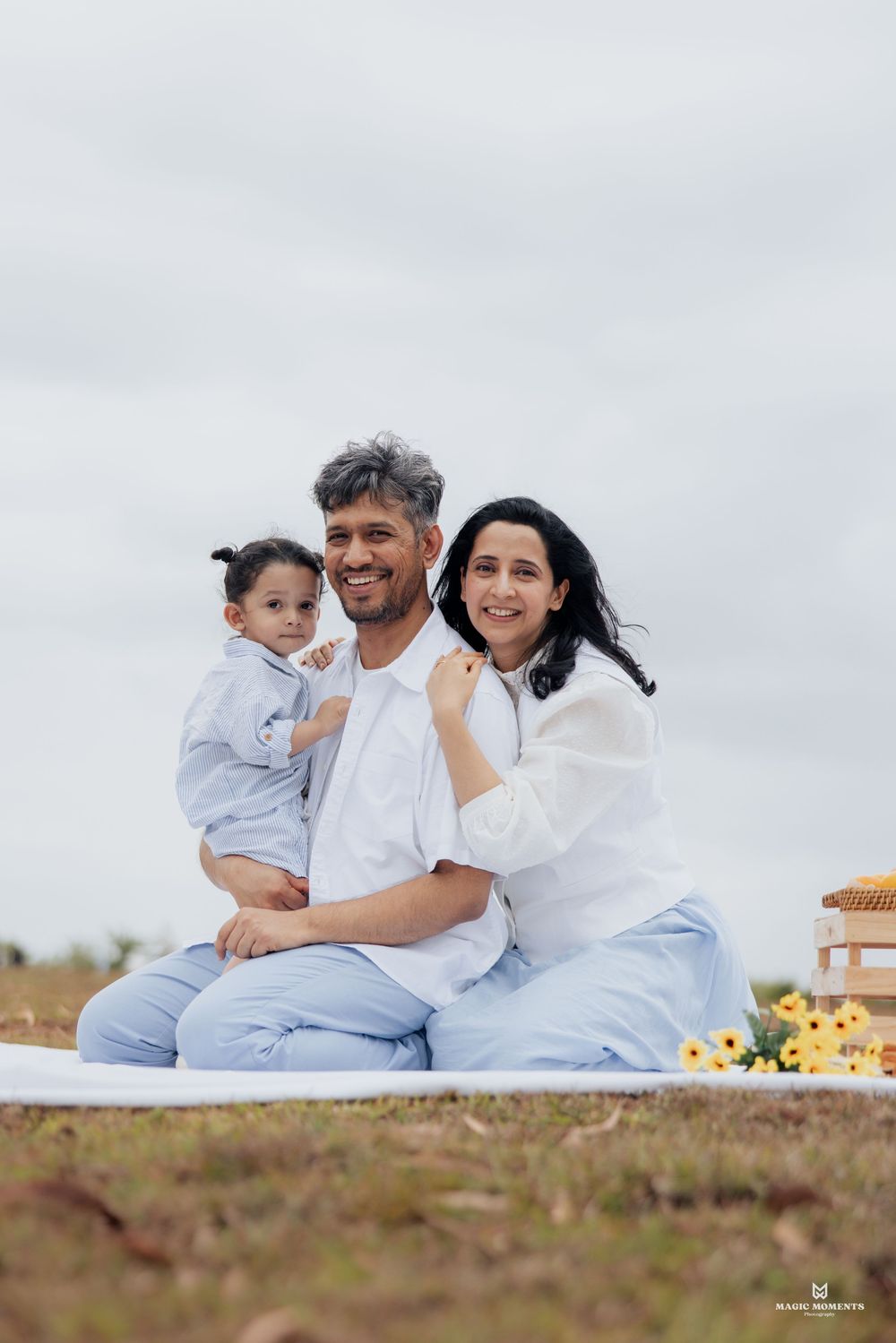 Happy family of three sitting on a white blanket outdoors, parents smiling with their toddler during a picnic photo session.