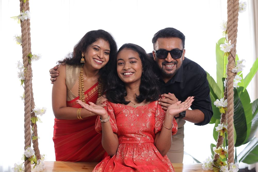 A joyful South Indian family of three posing on a swing, woman in red saree, girl in red dress, man in black shirt.