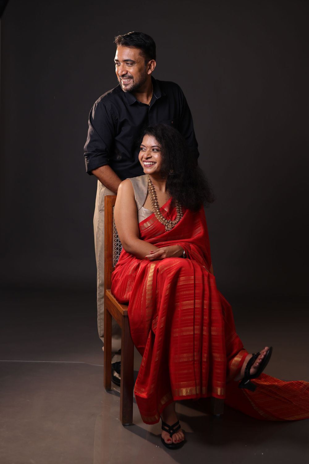 A couple posing in a studio, man in black shirt standing behind woman seated in red saree with gold jewelry.
