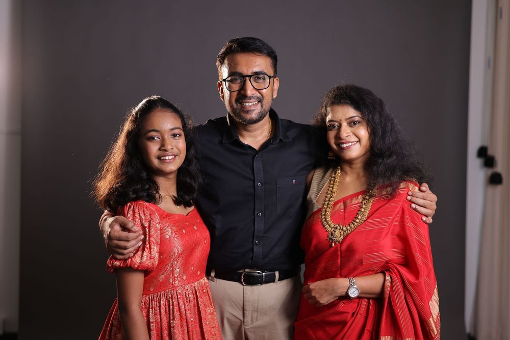 A smiling family of three posing together, man in black shirt with two women in red traditional Indian attire.