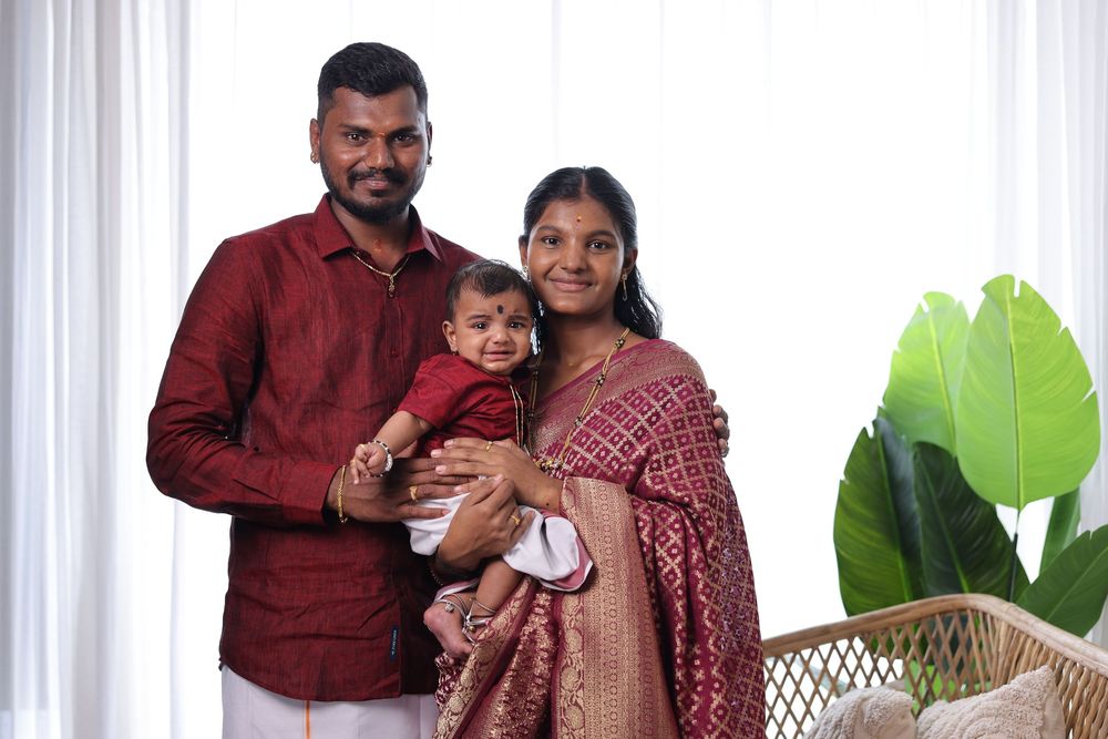 Indian family portrait with father, mother, and baby dressed in matching red traditional attire indoors.