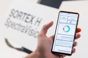 Hand holding smartphone displaying SORTEX H SpectraVision sorting machine analytics dashboard app.